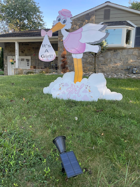 Decorative stork with baby bundle and 'It's a Girl!' text on a grassy lawn in front of a house.